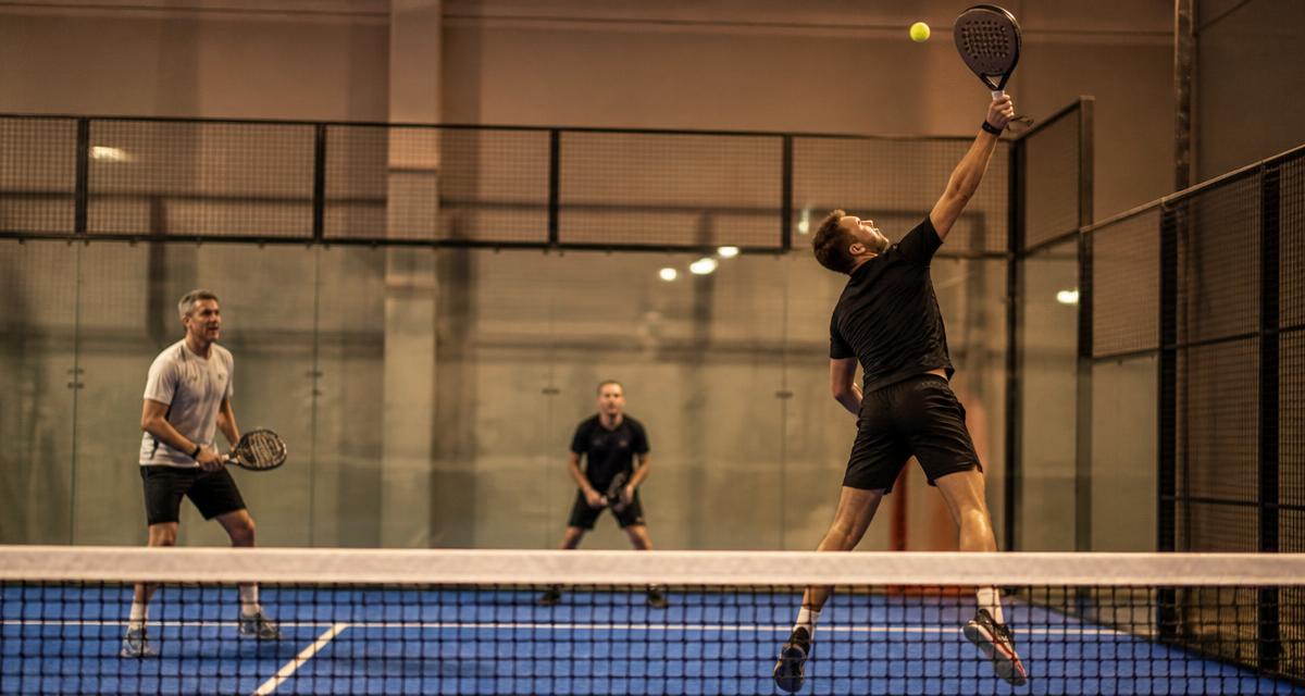 Padelspelare slår ett högt slag i axelhöjd framme vid nätet under en inomhusmatch i dubbel
