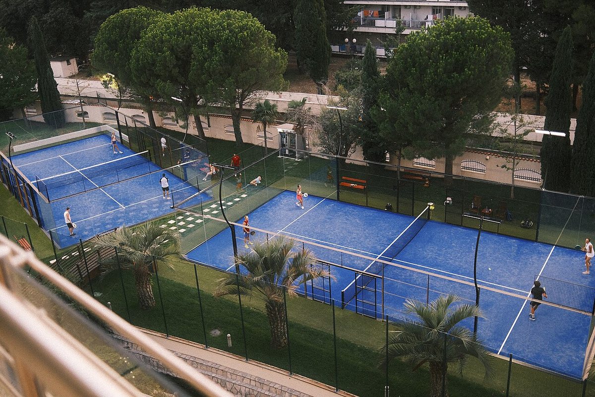 Two doubles matches on outdoor blue padel courts with glass walls and players on both sides of the nets